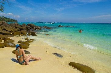 A girl in a bikini sits on white sand on the shore of the emerald sea