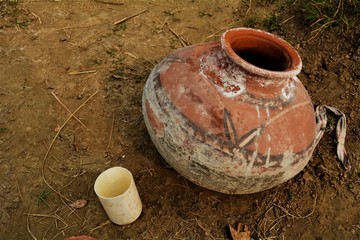 A simple handmade earthen pot used for water and a small jug sits in a field during a hot day.