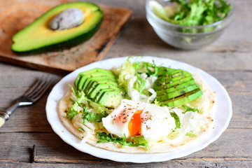 Healthy breakfast recipe. Homemade flour tortilla with a poached egg, avocado slices, napa cabbage, salad mix, sauce and spices on a plate. Avocado half, green salad in a glass bowl, fork on a table