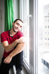 Young man smile to the camera in the morning sitting near window at his apparment