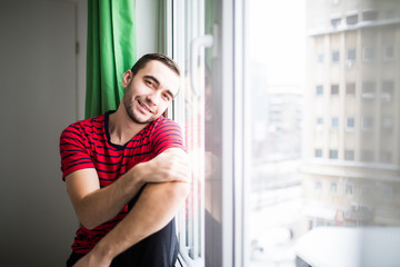 Young man smile to the camera in the morning sitting near window at his apparment