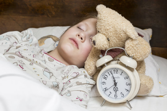 śpiące dziecko w ł&oacute;żku z misiem i budzikiem