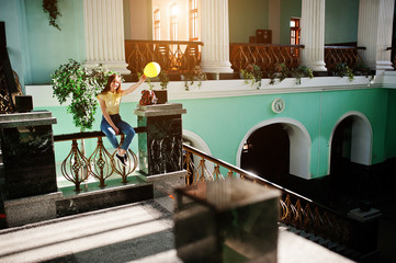 Young teenage girl sitting on stair railings at the station with ballon at hand, wear on yellow t-shirt, jeans and sunglasses.