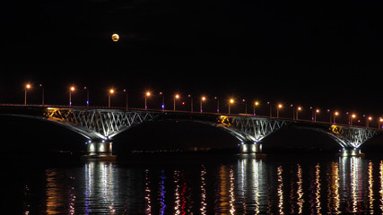 Naklejka na ściany i meble Full moon rises over the bridge. Road bridge between the cities of Saratov and Engels, Russia. The Volga River. The evening lights of cars and street lights