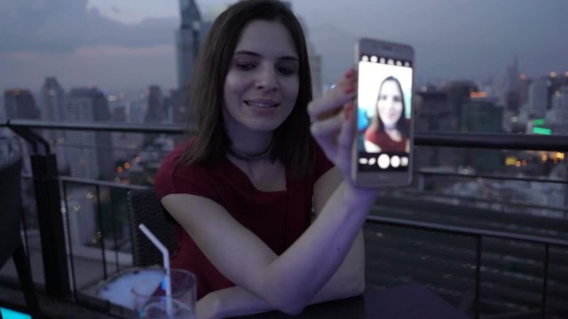 Young woman with cocktail taking selfie photo on terrace in bar at night
