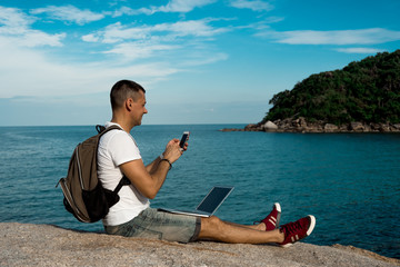 Man sits on a rock against the background of the sea with a smart phone and laptop  