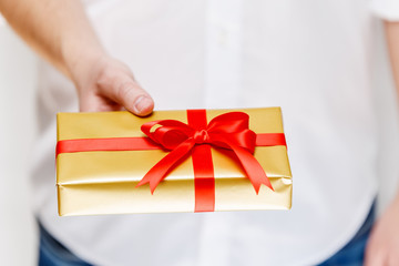 Male hand holding a gift box. Present wrapped with ribbon and bow. Christmas or birthday golden package. Man in white shirt.
