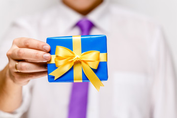 Male hand holding a gift box. Present wrapped with ribbon and bow. Christmas or birthday blue package. Man in white shirt and necktie.