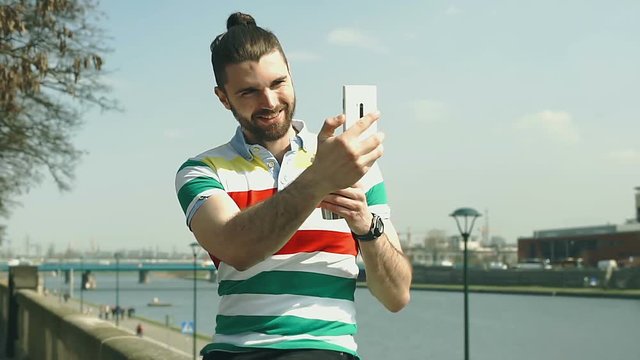 Handsome man with mud bun doing selfies on smartphone while standing next to the river
