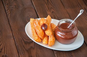 Churros sprinkled with sugar and cinnamon and hot chocolate in a glass jar on a white plate on a wooden background. Traditional spanish treat.  Copy Space.