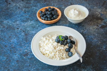 Organic Farming Cottage cheese in a white plate sour cream and blueberries on a blue stone background.