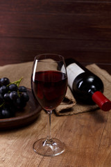 Red wine. Bottle with wineglass. Dark grapes branch on plate. On rustic wooden background.
