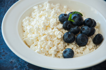 Organic Farming Cottage cheese in a white plate sour cream and blueberries on a blue stone background.