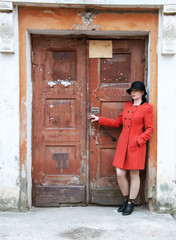 Young beautiful girl dressed in red coat posing near an old door from the entrance of the house. Female face in shadow.