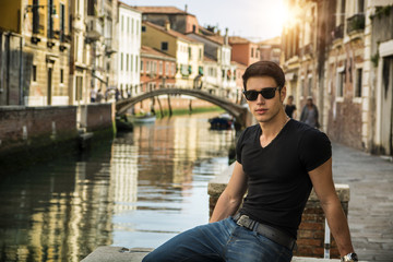 Fototapeta na wymiar Portrait of Attractive Dark Haired Young Man Leaning Against Railing on Foot Bridge Over Narrow Canal in Venice, Italy