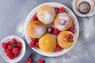 Homemade lemon muffin cupcakes with fresh raspberries, sugar powder, mint, served with vintage sieve over gray blue texture stone background. Top view with space.
