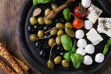 Mediterranean appetizer antipasti board with green black olives, feta cheese, mozzarella, capers, pepper, basil with grissini bread sticks over black wood burnt background. Top view with space