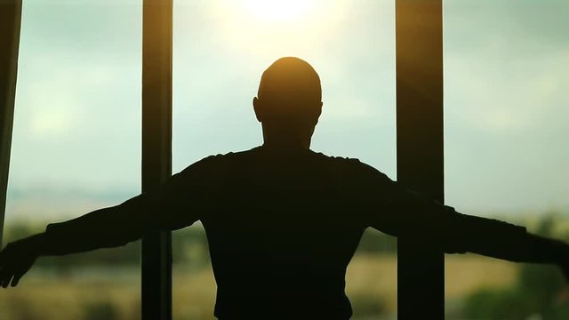 Man opens the curtains in the morning raises his hands and stretch oneself. Young man opens big window curtains and lets the sun light in the room