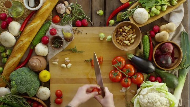 Top view of female hands cutting cherry tomatoes with knife on cutting board with variety of fresh produce on table. Footage in fast motion