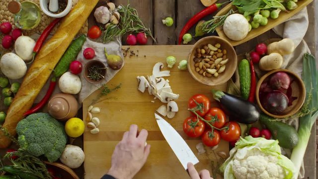 Top view of male hands slicing mushrooms with knife on wooden cutting board while cooking on table with lots of fresh vegetables on it. Fast motion shot