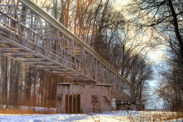 A winter photo of a metallic structure supporting various pipes on concrete blocks running through a forest with sun shining though the canopy