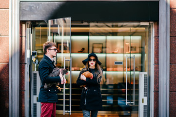 Fototapeta premium Man in black coat and opens door to a beautiful model long hair girl in glasses and elegant hat and fashion clothes coming out of boutique with chicken in hands after shopping. Celebrity pets concept.