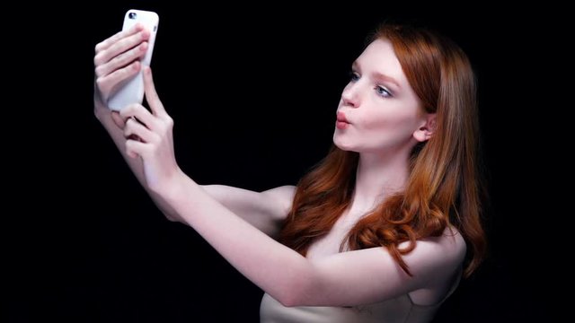 Young active woman with long red hair making selfie on mobile phone isolated over black