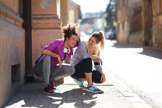 Two young woman taking a break after jogging ,early in the morning.They crouching and laughing.