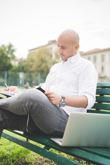 Fototapeta premium Young contemporary businessman remote working sitting on a bench outdoor in a city wiritng on her agenda - organizing, small business, remote working