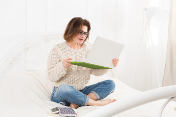 Young woman reads from laptop in bed, high key