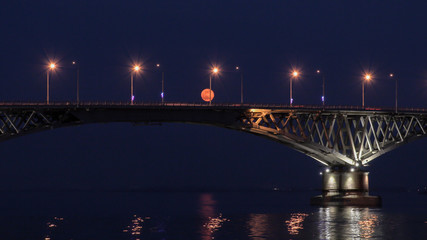 Fototapeta na wymiar Full moon rises over the bridge. Road bridge between the cities of Saratov and Engels, Russia. The Volga River. The evening lights of cars and street lights