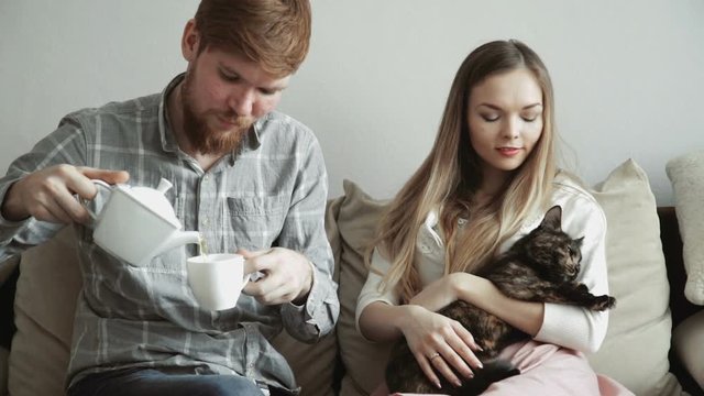 Couple is sitting on the couch sofa at home stroking a cat, Drink tea from a white tea set and watching tv.