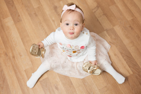 One Year Old Baby Girl Dressed In White Shirt And Satin Dress Sitting On The Floor Holding Golden Shoes In Her Hands; View From Above