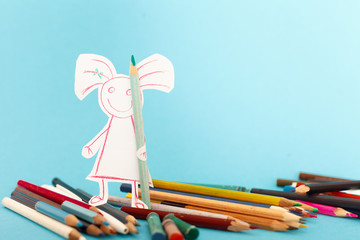 The paper doll of a girl stands among the colored pencils and holds one of them in her hands. Blue background. Children's creativity concept