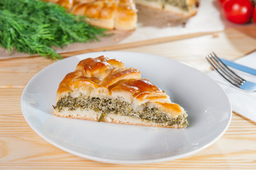 Fresh piece of vegetarian closed pie with Feta cheese and spinach on the white plate on the wooden served table. Composition of ingridients. Selective Focus, close up