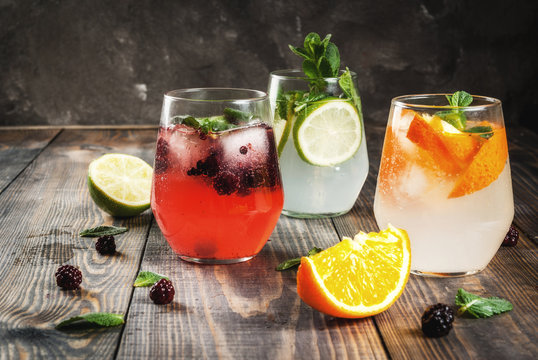Selection of three kinds of gin tonic: with blackberries, with orange, with lime and mint leaves. In glasses on a rustic wooden background. Copy space 