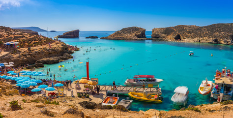 Comino, Malta - Tourists crowd at Blue Lagoon to enjoy the clear turquoise water on a sunny summer day with clear blue sky and boats on Comino island, Malta.
