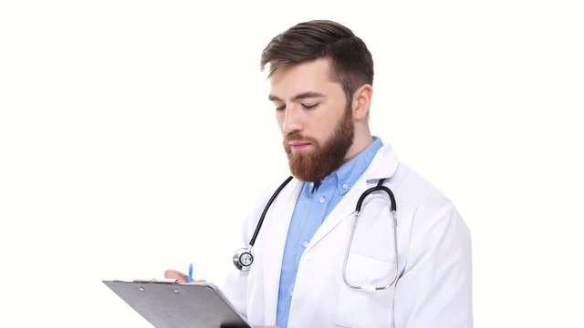 Serious smiling young male doctor listening and writing to folder information isolated