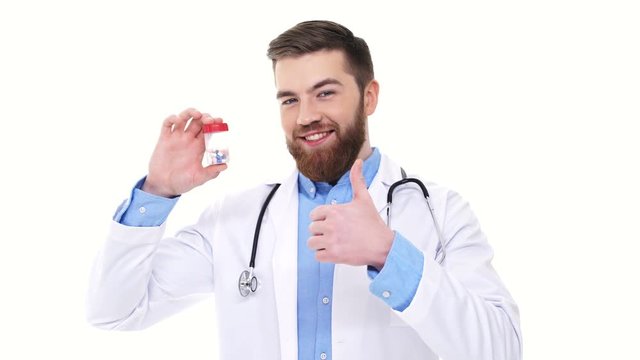 Smiling male doctor winking while showing bottle with pills and gesture thumb up isolated