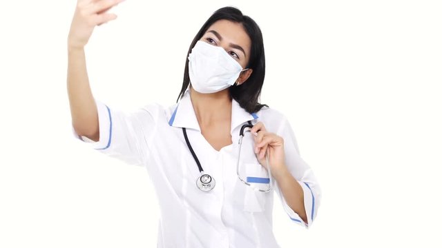 Woman nurse wearing white face mask making selfie using smartphone and showing gesture two fingers isolated
