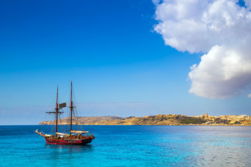 Fototapeta na wymiar Blue Lagoon, Malta - Old sailing boat at the Island of Comino next to the famous Blue Lagoon with the Island of Gozo and town of Mgarr at the background on a sunny summer day