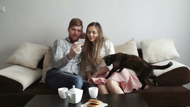Couple is sitting on the couch sofa at home stroking a cat, Drink tea from a white tea set and watching tv.