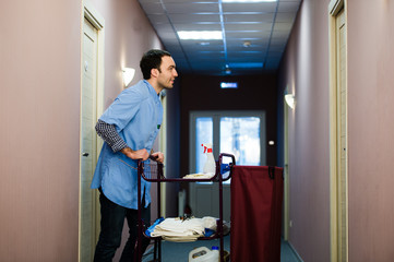 Young man pushing a housekeeping cart laden with clean towels, laundry and cleaning equipment in a hotel as he services the rooms