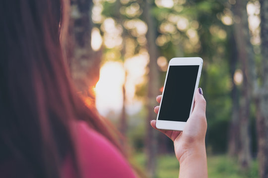 Mockup image of a woman holding and using smart phone with blank black screen at outdoor and green nature background during sunset time