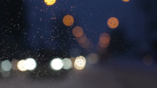 Snowy evening at city. Water drops on windshield glass window, car lights out of focus background