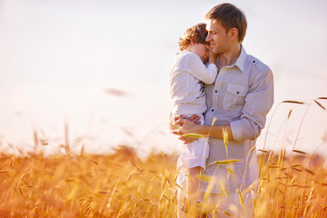 A man is holding his son in his arms, standing in the middle of a wheat field. The son gently laid his head on his father's shoulder, hugging him