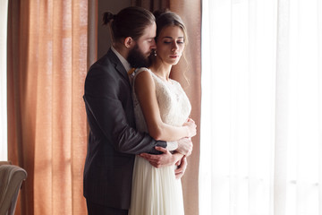 The bridegroom is hugging the bride from the back, standing by the window. Side view