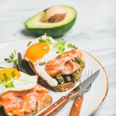 Healthy breakfast sandwiches. Salmon, avocado, fried egg, sauted green beans and fresh sprouts sandwiches in white plate over marble background, selective focus, square crop. Clean eating food concept