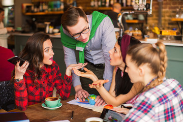 Group of students sits at the table with cups of coffee and working things and discusses about their affairs in time of a break in a cafe