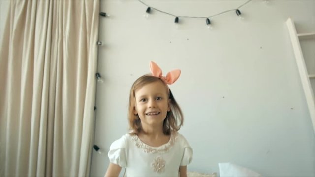 A happy cute young girl in white dress having fun jumping on bed, little smiling child